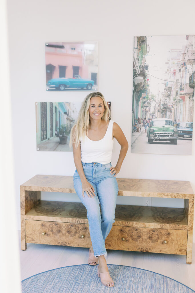 A smiling woman sitting on a wooden bench in a brightly lit room, surrounded by colorful photographs of vintage cars and vibrant streets.